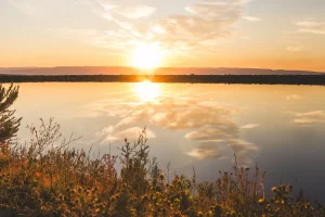 Sunset over a calm body of water with vibrant orange and pink hues reflecting on the surface, surrounded by vegetation, illustrating the natural beauty of water sources essential to Idaho water rights.