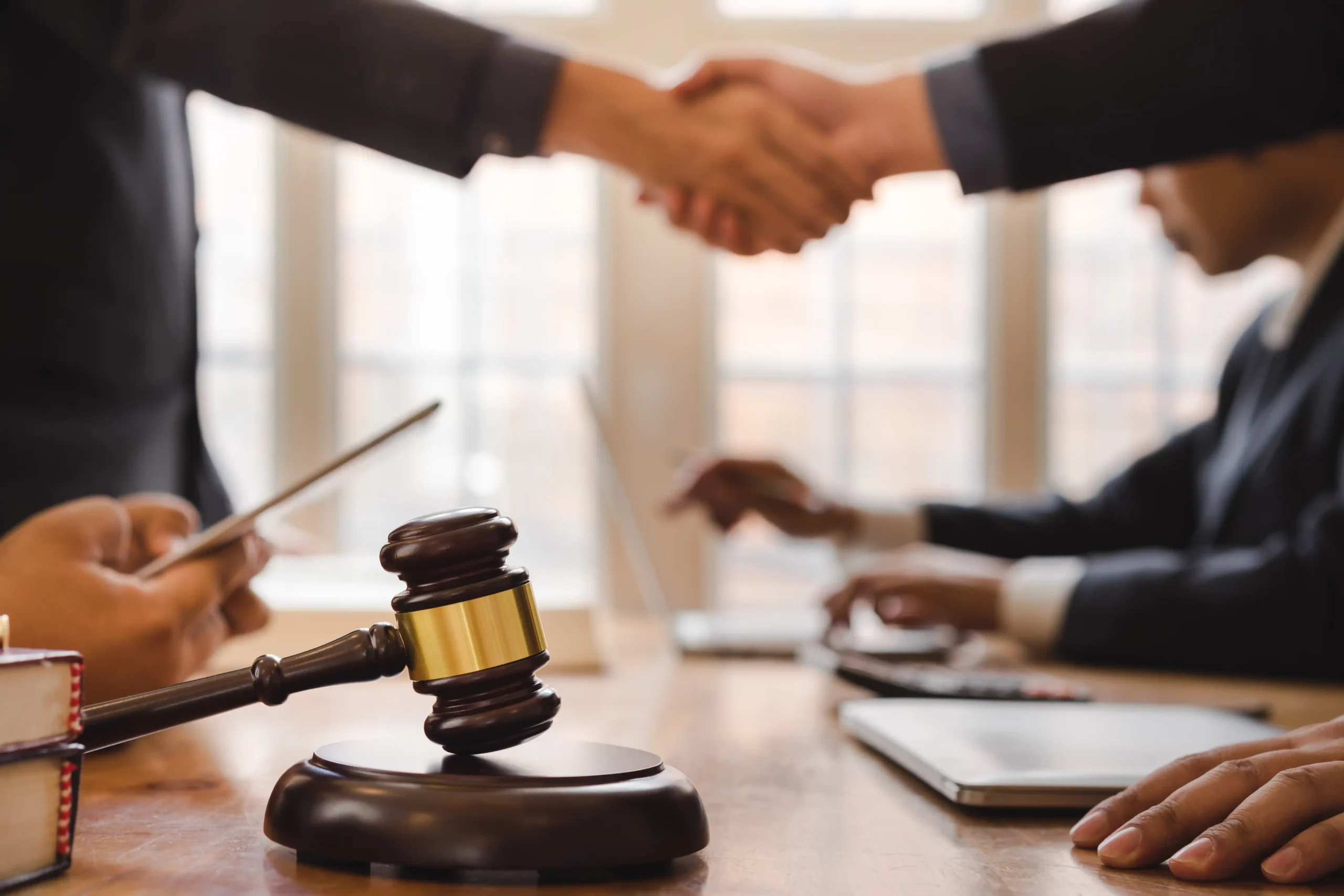 This image shows a close-up of a gavel resting on a table, symbolizing a legal setting, while two individuals in business attire shake hands in the background. The scene conveys a sense of agreement or negotiation, with other participants focusing on documents and technology around them. The image suggests a legal or professional context, possibly related to water rights in Idaho, with the handshake representing the resolution of a legal matter or contract involving these rights.
